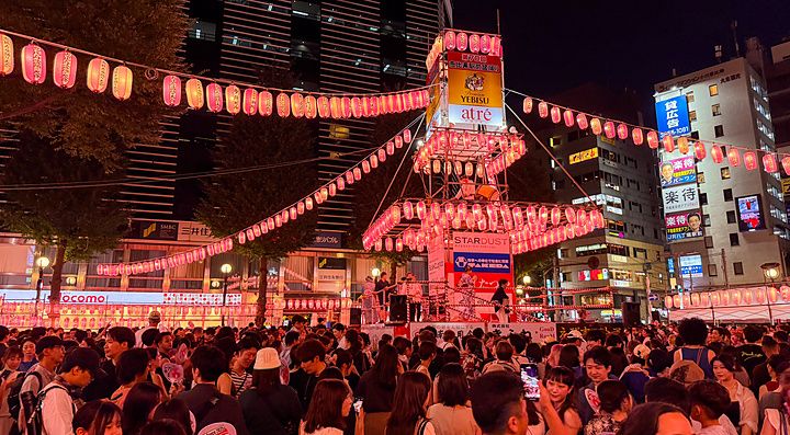 Una folla di persone partecipa a un matsuri ad Ebisu davanti alla stazione, con un palco centrale illuminato da lanterne.