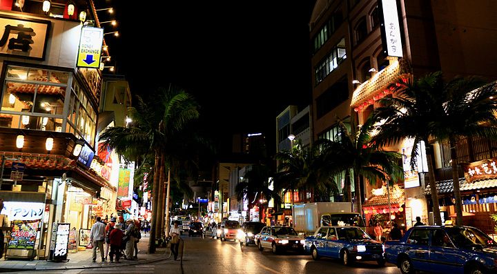 Veduta notturna di Kokusai Street a Naha, Okinawa, con insegne luminose, palme e traffico cittadino.