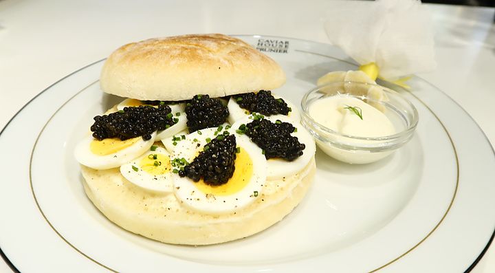 Un panino di lusso con caviale e uova sode su un piatto con logo Caviar House & Prunier accanto a una salsa bianca.
