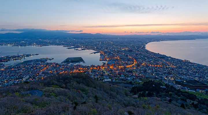 Veduta aerea notturna di Hakodate con luci cittadine e mare ai lati all'alba.
