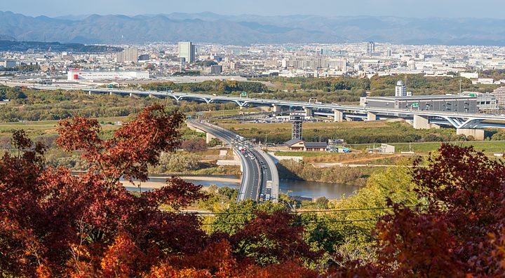 Veduta panoramica di Yawata City e le montagne sullo sfondo, incorniciata da alberi con foglie autunnali dai toni caldi.