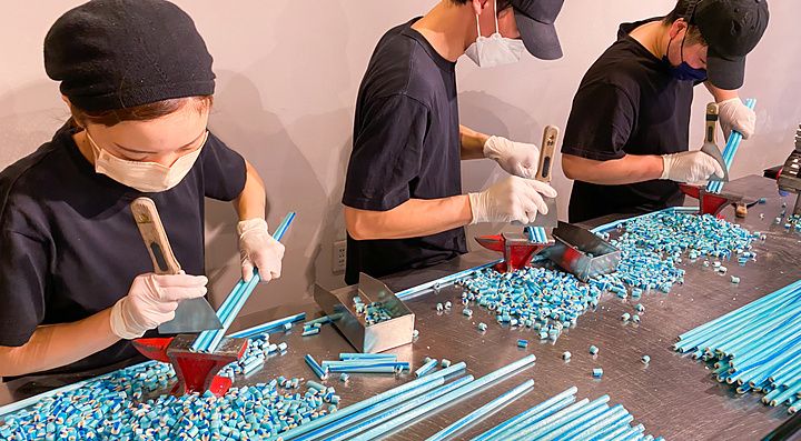 Tre persone con maschere e guanti tagliano lunghi bastoncini di caramelle artigianali azzurre su un tavolo in acciaio.
