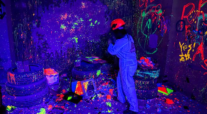 Una persona con tuta e casco distrugge oggetti nella rage room a Reeast Room, in un ambiente illuminato da luci UV.