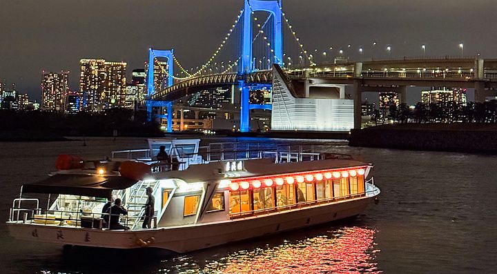 Veduta notturna di una Yakatabune che naviga vicino ad Odaiba, con il Rainbow Bridge illuminato sullo sfondo.