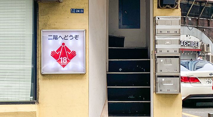 L'ingresso di un locale per adulti a Shimbashi con insegna luminosa, una rampa di scale e pareti color crema.