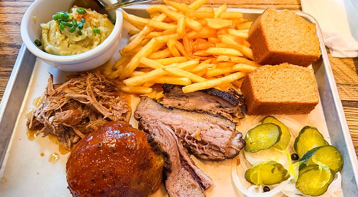 Un vassoio metallico di Smokehouse con barbecue, patatine fritte, pane di mais, purè e cetriolini.