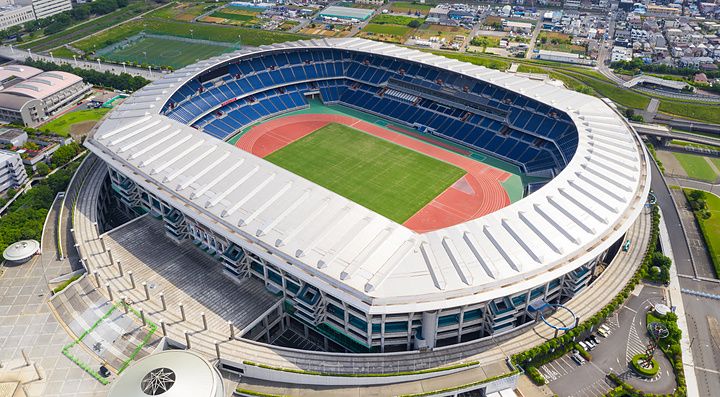 Veduta aerea del Nissan Stadium di Yokohama con il campo centrale circondato da tribune blu e una pista rossa.