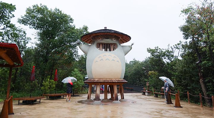 Grande statua in legno di Totoro in un parco boschivo sotto una pioggia leggera con visitatori.