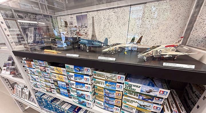 Scaffali pieni di scatole di modellini Tamiya sotto una vetrina con aerei assemblati al Tamiya PlaModel Factory a Tokyo.