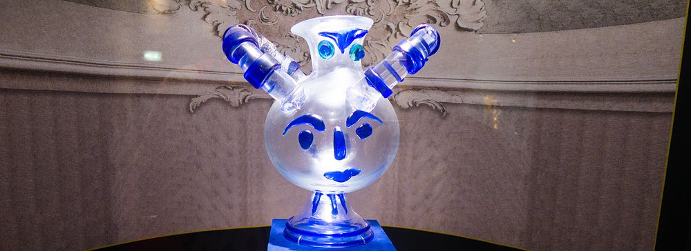 Un'anfora in vetro veneziano con decorazioni a forma di volto esposta su un piedistallo blu al Hakone Glass Forest Museum.