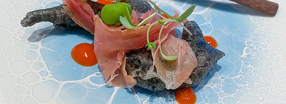 Un antipasto al Koji Shimomura composto da gnocco fritto con calamaro e prosciutto crudo italiano su un piatto azzurro.