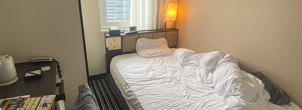 Interno di una camera compatta presso l'Apa Hotel a Shinjuku con letto singolo disfatto e finestra urbana.
