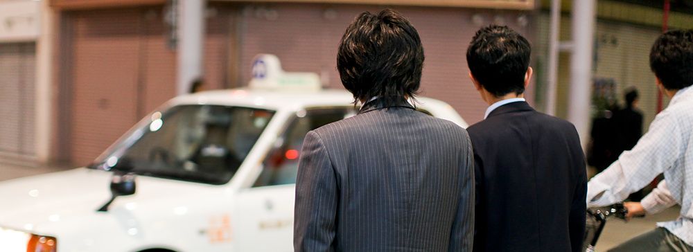 Due persone in abito formale attendono un taxi bianco in una strada urbana in Giappone.