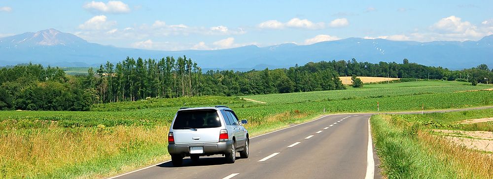 Un'auto grigia viaggia lungo una strada asfaltata tra campi verdi e montagne in una giornata serena a Biei, Hokkaido.