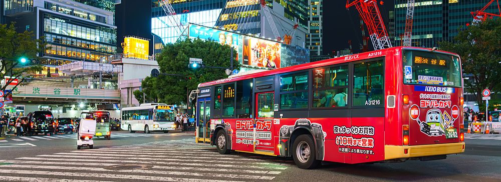 Veduta notturna dell'incrocio di Shibuya a Tokyo con un autobus rosso che attraversa la strada tra i grattacieli.