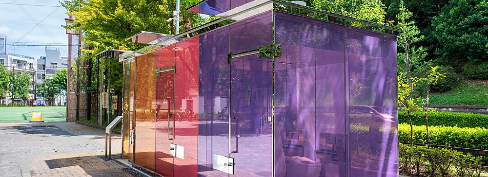 Struttura di un bagno pubblico moderno con pareti in vetro colorato arancione, rosso e viola situata in un parco urbano.