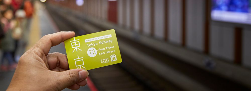 Una mano tiene un biglietto verde per la Tokyo Subway su una banchina ferroviaria sfocata.