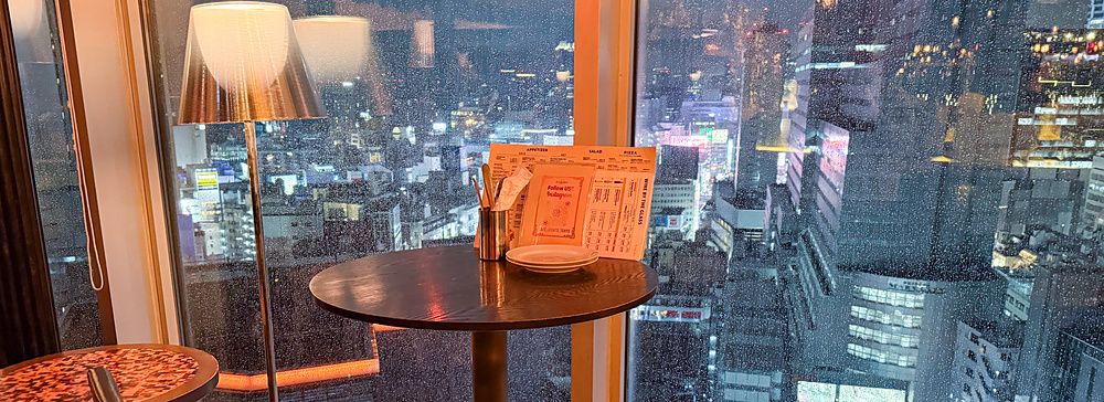 Tavoli e sedie di Cafe Legato a Shibuya posizionati accanto a una grande vetrata che affaccia sulla città piovosa.