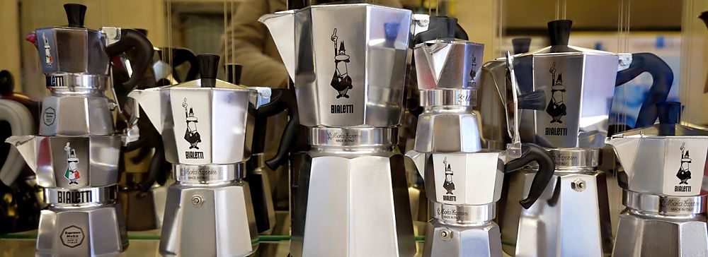 Caffettiere Bialetti Moka Express in alluminio di varie dimensioni disposte su un ripiano in vetro in un negozio.