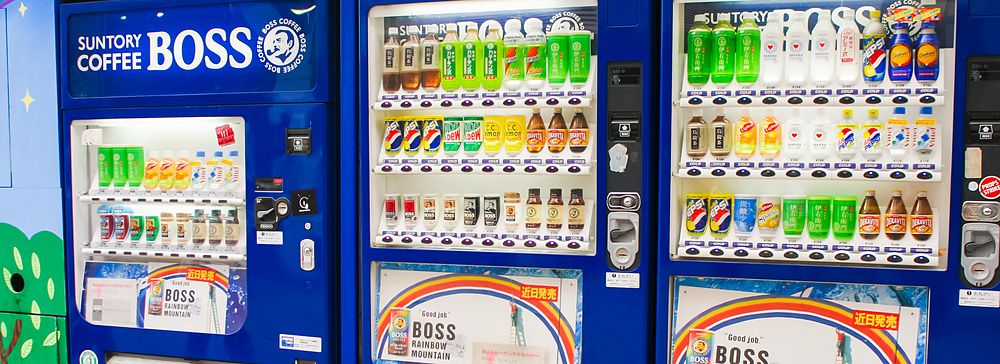 Tre distributori automatici di bevande Suntory Boss blu allineati su una strada in Giappone.
