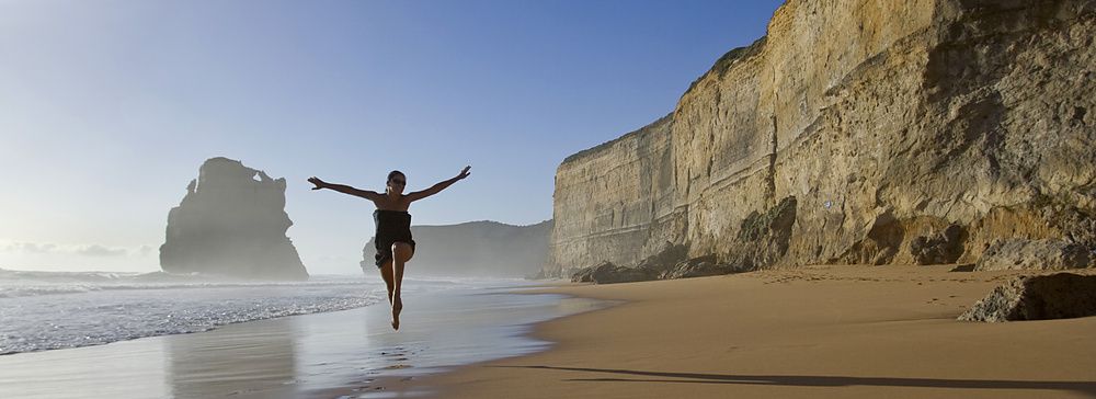 Donna felice che salta sulla spiaggia accanto a scogliere imponenti.