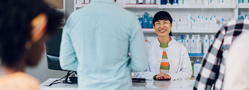 Una farmacista sorridente parla con un cliente al bancone di una farmacia ben fornita.
