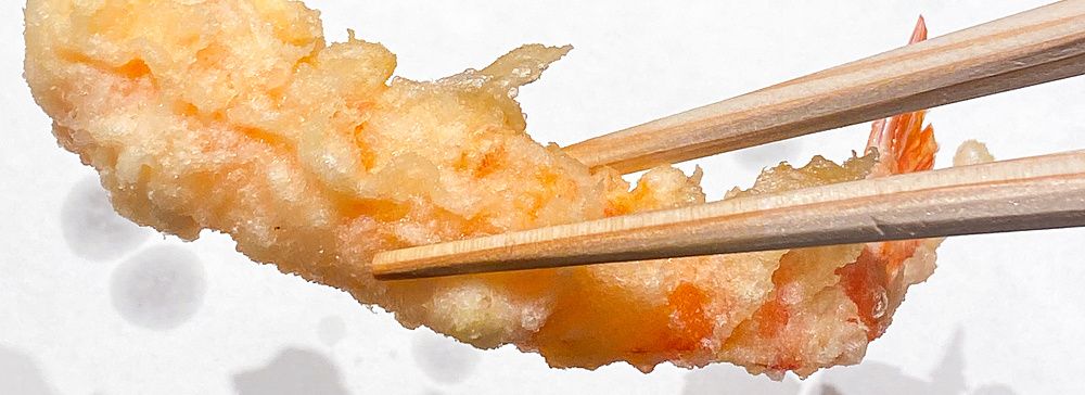 Un gambero a Tempura Kondo sollevato da un paio di bacchette di legno sopra un vassoio coperto di carta.