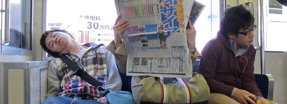 Tre passeggeri siedono su un treno pendolari in Giappone, con uno dei soggetti che dorme e un altro che legge il giornale.