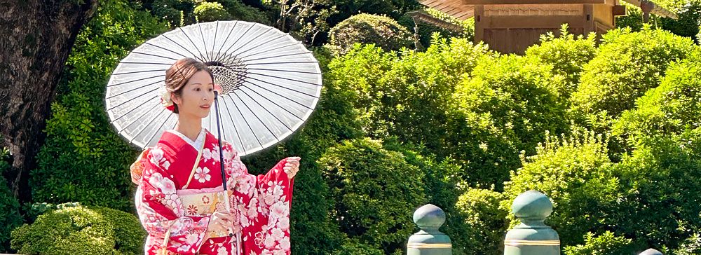 Una donna in kimono rosso tiene un ombrello bianco su un ponte in un giardino a Tokyo.
