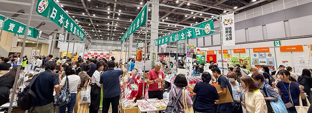 Interno della Good Life Fair a Tokyo con numerosi stand espositivi, visitatori e insegne in giapponese nel padiglione.