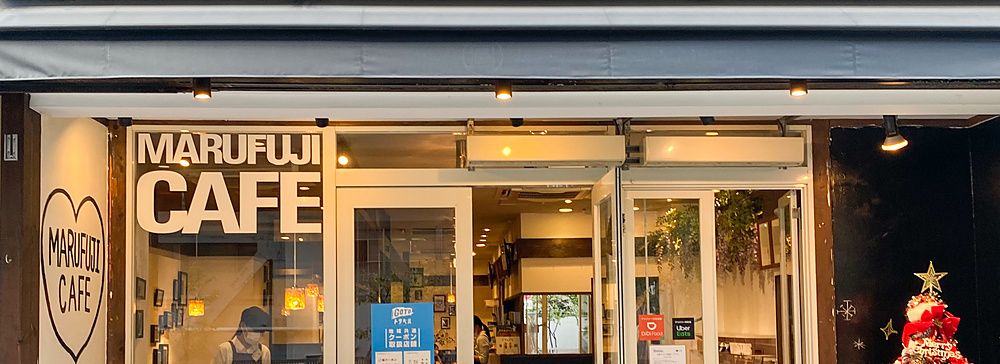 L'ingresso a Marufuji, un caffè con insegna bianca, vetrate, albero di Natale all'esterno e persone all'interno.