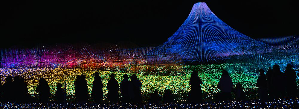 Sagome di visitatori che osservano una grande installazione luminosa a forma di monte Fuji durante un festival notturno.