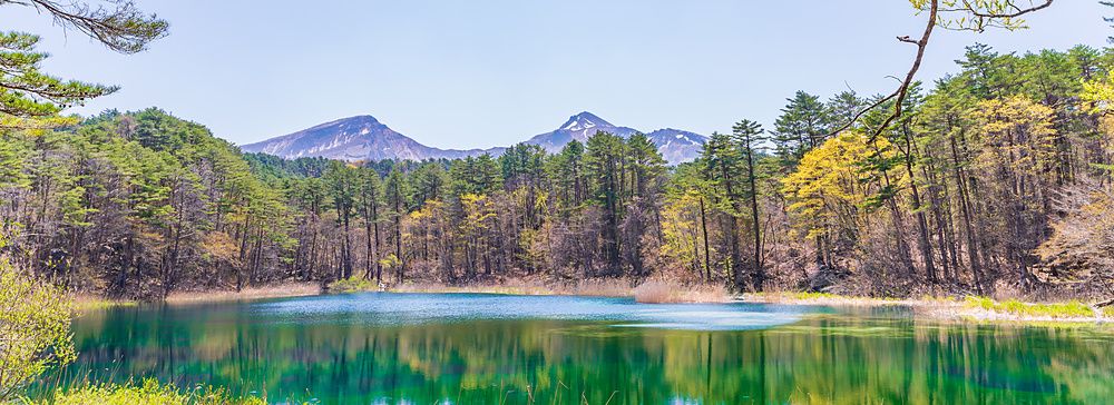 Veduta panoramica del Ruri-numa a Goshikinuma con montagne innevate sullo sfondo e riflessi boschivi nell'acqua turchese.