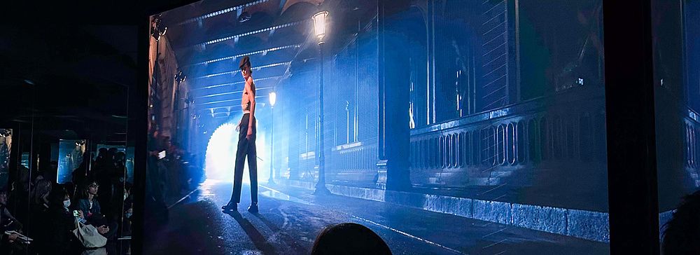 Una persona osserva una grande proiezione cinematografica di una figura maschile in una strada notturna alla mostra di Mai...