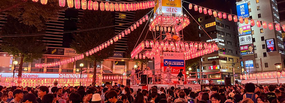 Una folla di persone partecipa a un matsuri ad Ebisu davanti alla stazione, con un palco centrale illuminato da lanterne.