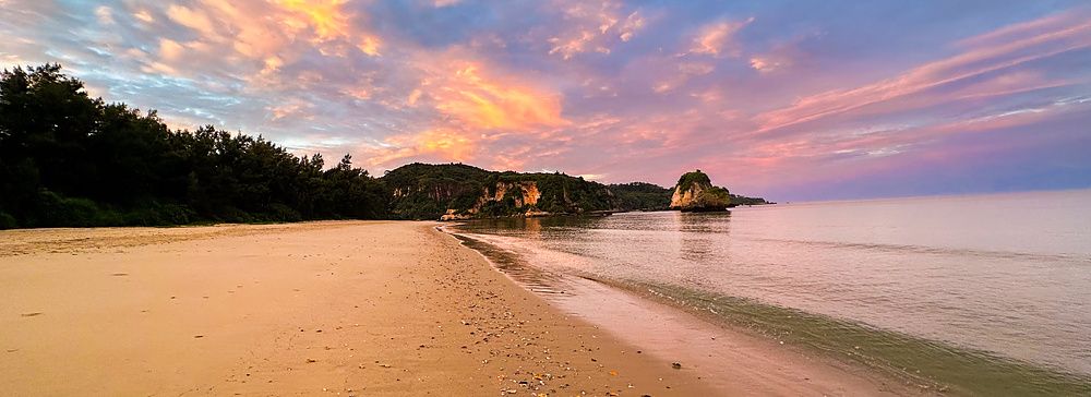 Una veduta della spiaggia Mimikiri Beach a Okinawa con sabbia dorata, vegetazione rigogliosa e un cielo al tramonto.