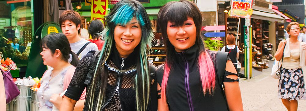 Due persone sorridenti vestite in modo alternativo camminano a Shibuya tra la folla.