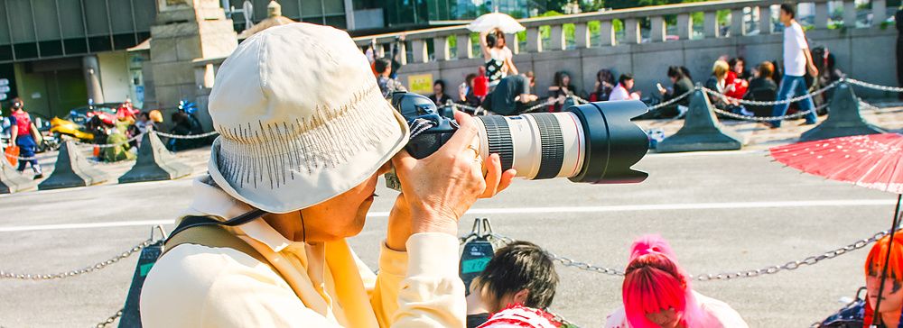 Una persona con cappello chiaro fotografa individui in costume ad Harajuku durante una giornata di sole.