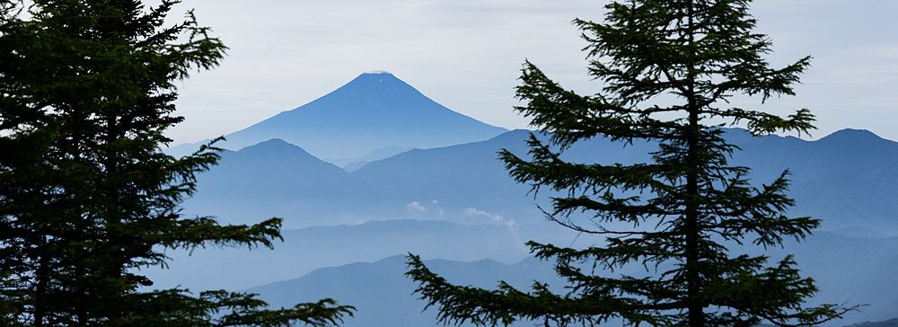 Veduta del monte Fuji oltre strati di cime montuose, incorniciato da rami di pino dal monte Kumotori.