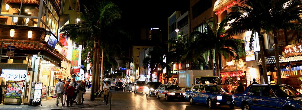 Veduta notturna di Kokusai Street a Naha, Okinawa, con insegne luminose, palme e traffico cittadino.