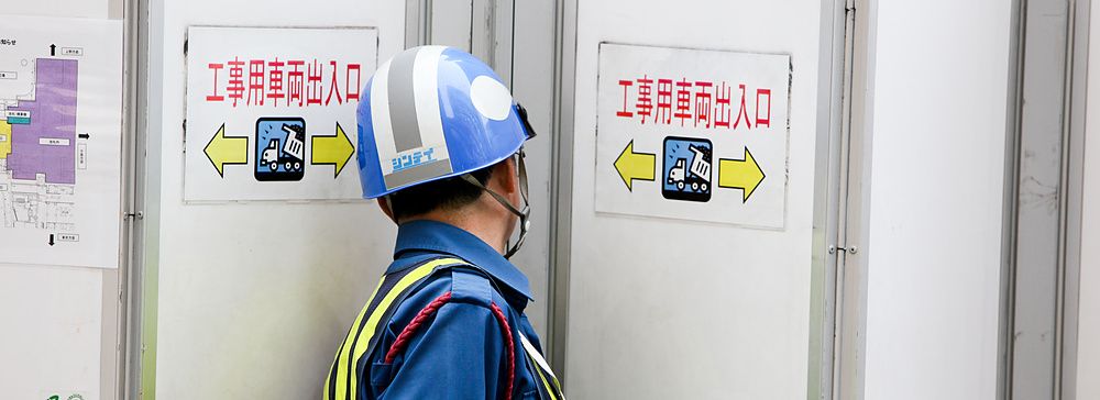 Un operaio giapponese con elmetto davanti a barriere e cartelli.