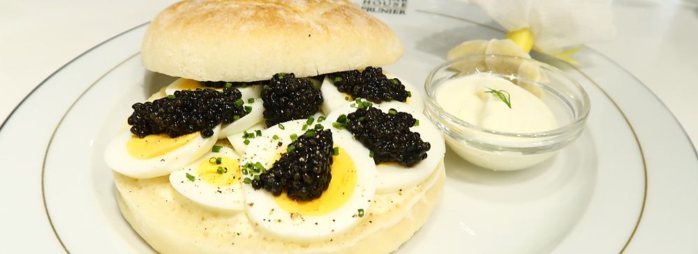 Un panino di lusso con caviale e uova sode su un piatto con logo Caviar House & Prunier accanto a una salsa bianca.
