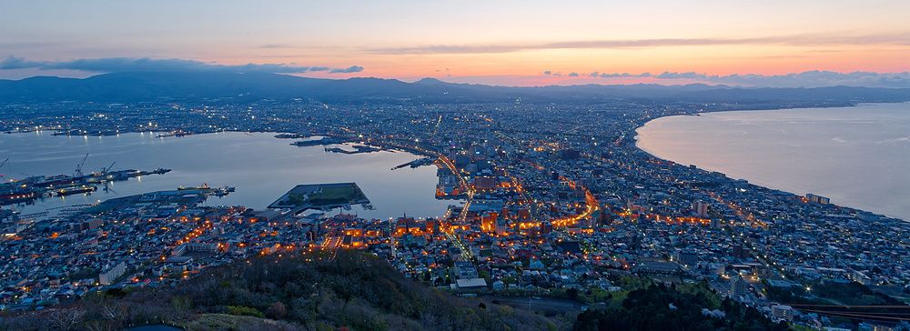 Veduta aerea notturna di Hakodate con luci cittadine e mare ai lati all'alba.