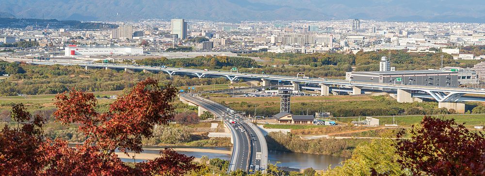 Veduta panoramica di Yawata City e le montagne sullo sfondo, incorniciata da alberi con foglie autunnali dai toni caldi.
