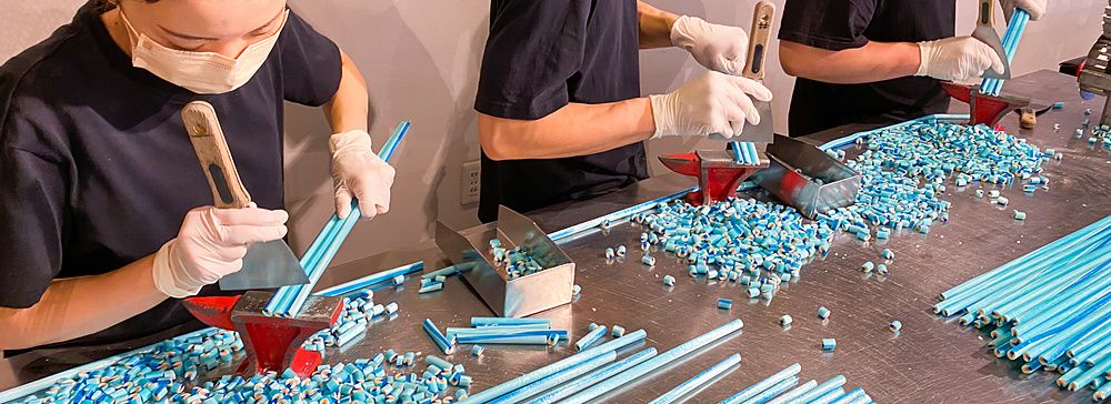 Tre persone con maschere e guanti tagliano lunghi bastoncini di caramelle artigianali azzurre su un tavolo in acciaio.