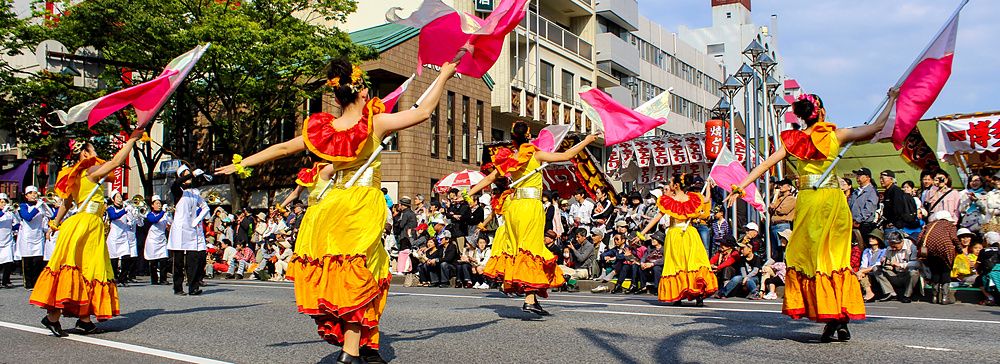 Danzatrici in abiti giallo-rosso sventolano bandiere colorate durante una parata stradale a Fukuoka.