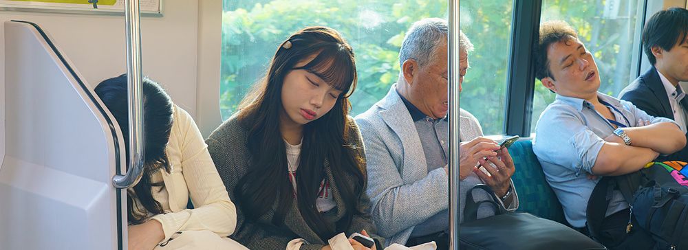 Passeggeri seduti su un treno a Tokyo, alcuni dormono e uno utilizza lo smartphone.