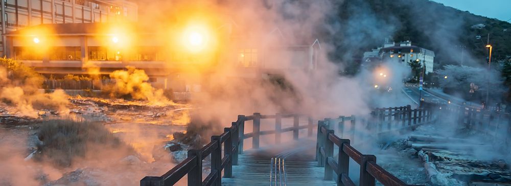 Passerella di legno che attraversa una valle geotermica fumante di notte con un hotel illuminato sullo sfondo.