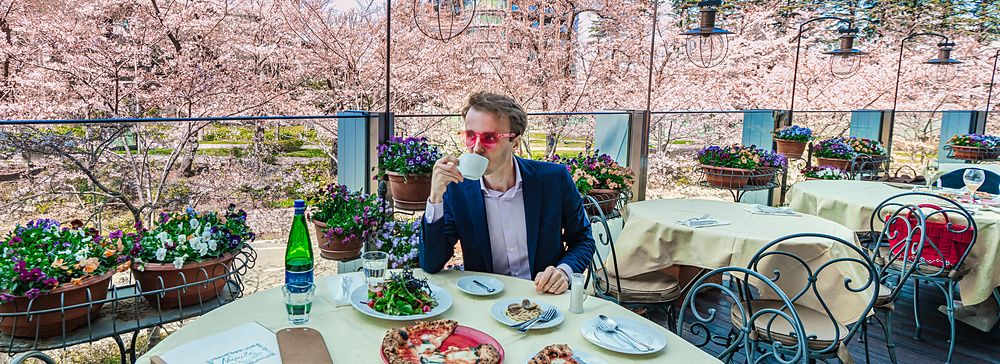 Marco Togni siede a un tavolo su una terrazza a Roppongi, circondato da ciliegi in fiore con un grattacielo sullo sfondo.