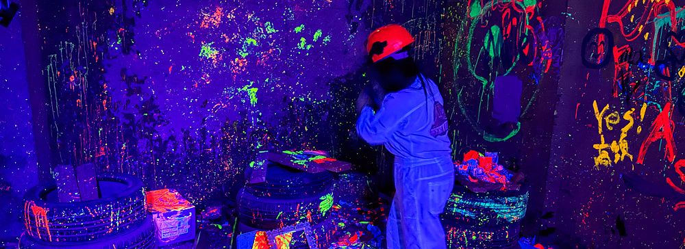 Una persona con tuta e casco distrugge oggetti nella rage room a Reeast Room, in un ambiente illuminato da luci UV.